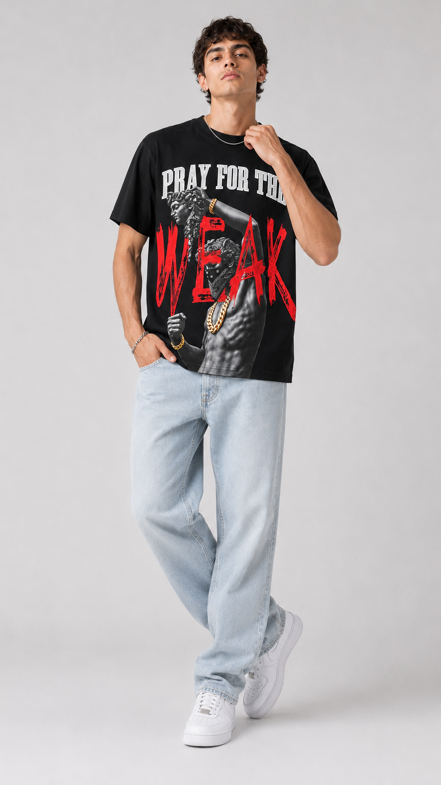 A man modeling a black graphic t-shirt with the text 'PRAY FOR THE WEAK' in white and red, paired with light-wash jeans and white sneakers, against a light gray studio background.