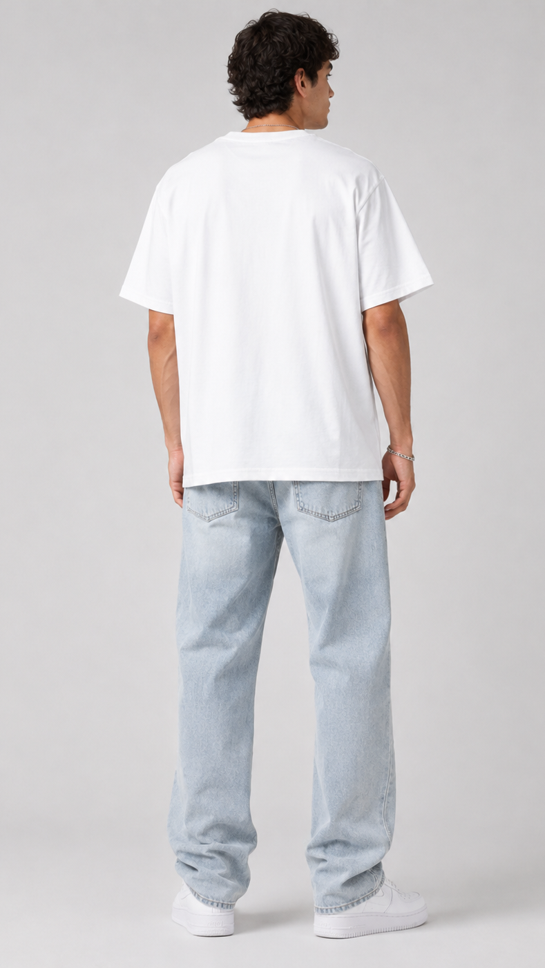 A man viewed from behind wearing an oversized white t-shirt, light-wash blue jeans, and white sneakers, standing against a neutral light gray studio background.