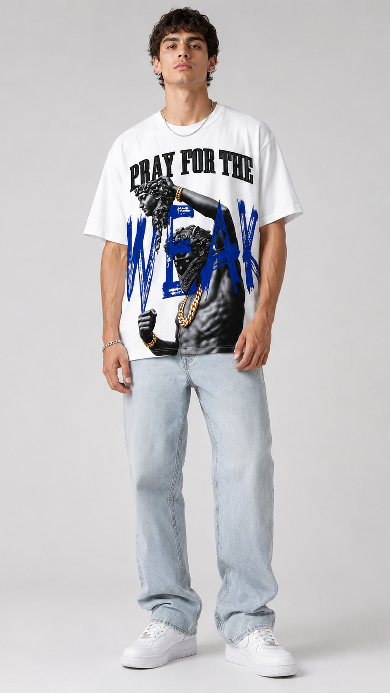 A male model stands against a light gray studio background wearing a white graphic T-shirt with black and blue text and imagery, light-wash jeans, and white sneakers.
