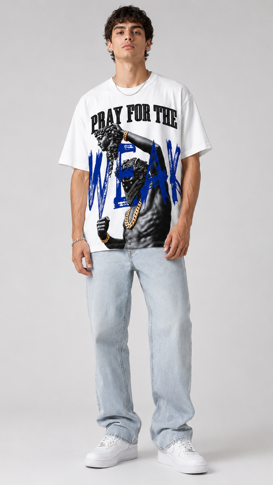A male model stands against a light gray studio background wearing a white graphic T-shirt with black and blue text and imagery, light-wash jeans, and white sneakers.