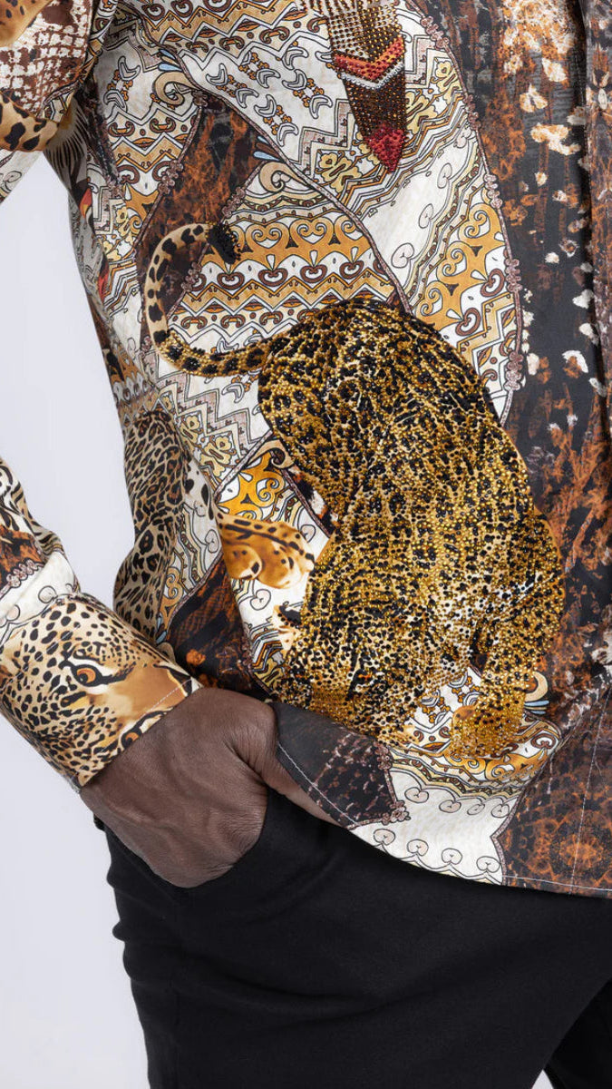 A close-up of a men's long-sleeve button-up shirt featuring a complex, multi-patterned print with leopard, cheetah, snake, and tribal motifs in brown, gold, black, and white. The shirt is shown on a model with a hand in their pocket, paired with black trousers.