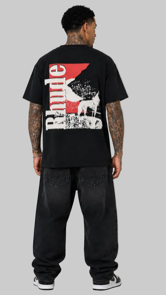 A man viewed from behind wearing a black t-shirt with a large red and white graphic print on the back, paired with baggy black jeans and white sneakers, set against a plain light gray studio background.