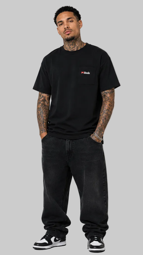 A man modeling a black short-sleeve t-shirt with a small red and white  logo on the left chest, paired with black jeans and black-and-white sneakers, standing against a neutral light gray studio background.