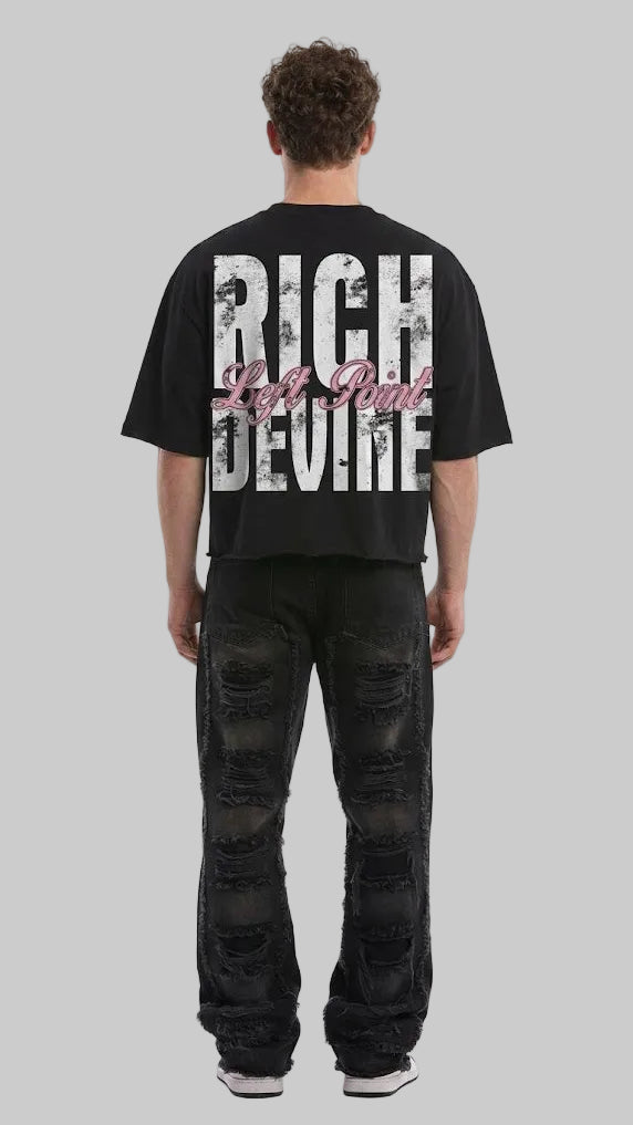 A man viewed from behind wearing a black oversized t-shirt with a large distressed white graphic reading 'RICH DEVINE' and pink script text, paired with distressed black jeans, against a light gray studio background.