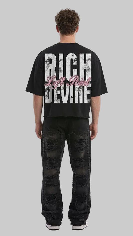 A man viewed from behind wearing a black oversized t-shirt with a large distressed white graphic reading 'RICH DEVINE' and pink script text, paired with distressed black jeans, against a light gray studio background.