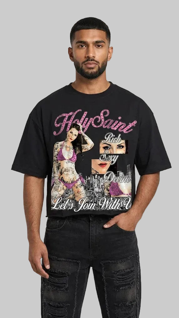 A man wearing a black graphic t-shirt with pink and white text reading 'HolySaint Rich Sexy Queen' and a collage print, paired with black jeans, against a light gray background.