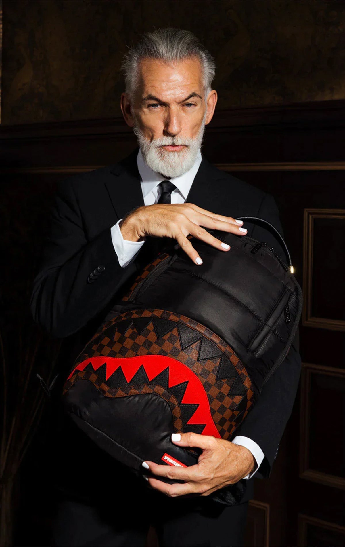 Black Sprayground backpack with a horizontal quilted puffer jacket-inspired design. Features multiple compartments including a separate velour laptop compartment, side pockets, and a hidden zippered pocket.  Equipped with ergonomic mesh back padding, adjustable straps, and a sliding back sleeve for attaching to carry-on luggage. Made from durable 100% polyester techy fabric with horizontal quilting.