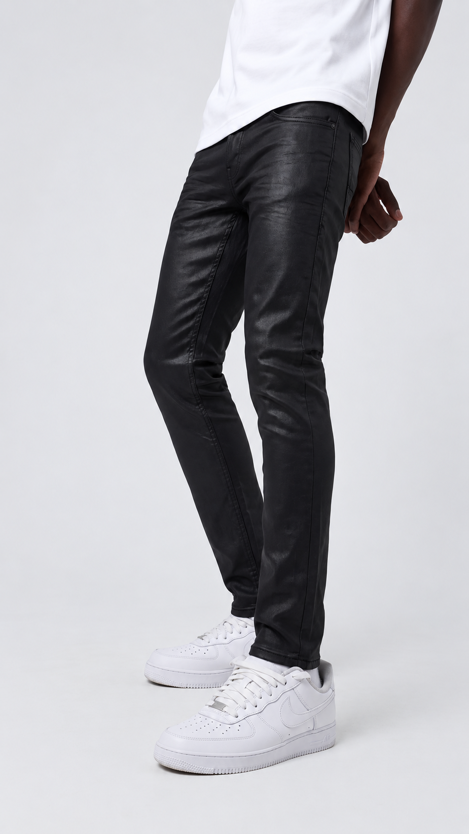 A person wearing slim black leather-look jeans with a white t-shirt and white sneakers, shown from the waist down against a light gray studio background.